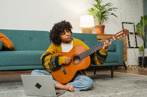 Person learning the guitar Person sitzt auf dem Boden im Schneidersitz mit einer Gitarre auf dem Schoß. vor der Person steht ein Laptop und sie sitzt in einem Wohnzimmer.