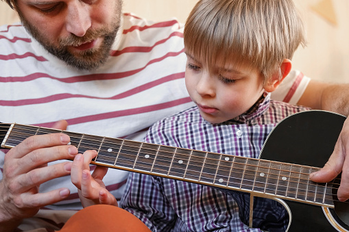 Man teaches child how to play the guitar Mann mit einem Kind auf dem Schoß. Bringt dem Kind Griffe auf der Gitarre, welche vom Mann vor dem Kind gehalten wird, bei.