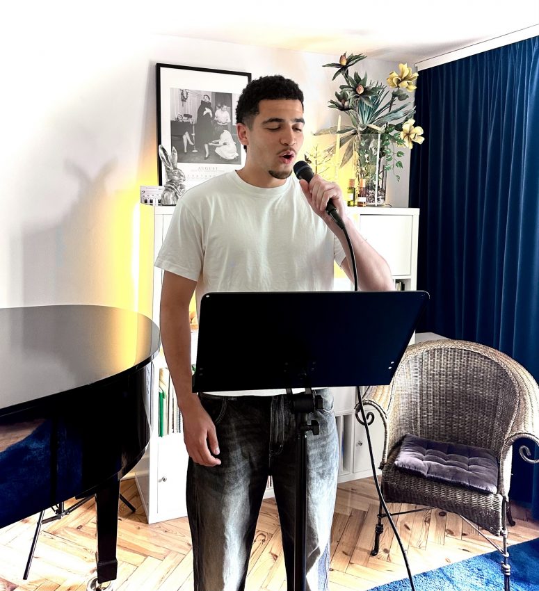 Mann steht vor einem schwarzen Notenpult. Er hat ein Mikrofon in der Hand und singt.